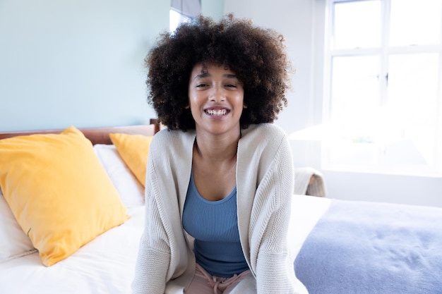 Foto porträt einer lächelnden gemischtrassigen jungen frau mit afro-haaren und jacke, die es sich zu hause im bett gemütlich macht. fröhliches, unverändertes lebensstil- und wohnkonzept.