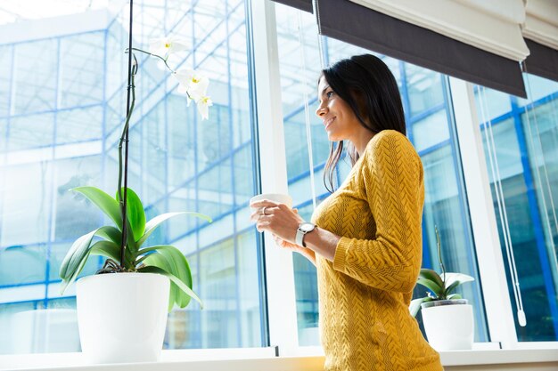 Porträt einer lächelnden Geschäftsfrau, die Tasse mit Kaffee hält und Fenster im Büro betrachtet