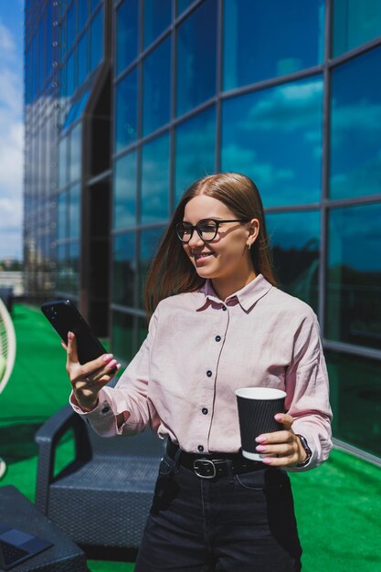 Porträt einer lächelnden Geschäftsfrau, die während der Kaffeepause auf dem Bürobalkon SMS auf dem Smartphone liest Moderne Terrasse im Büro zum Entspannen