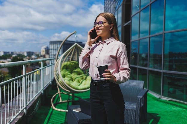 Porträt einer lächelnden Geschäftsfrau, die während der Kaffeepause auf dem Bürobalkon SMS auf dem Smartphone liest Moderne Terrasse im Büro zum Entspannen