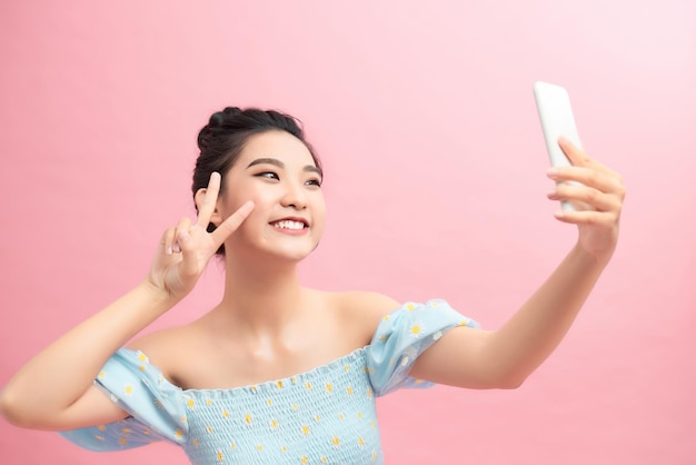 Porträt einer lächelnden, glücklichen Frau, die Victory-Zeichen zeigt und das Handy einzeln auf rosafarbenem Hintergrund betrachtet