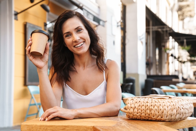 Porträt einer lächelnden glücklichen Frau mit Strohsack, die Kaffee aus Pappbecher trinkt, während sie im gemütlichen Café im Freien sitzt