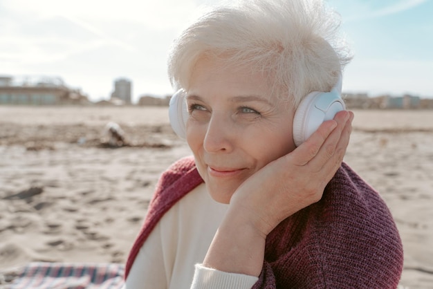 Porträt einer lächelnden, glücklichen, schönen Seniorin mit drahtlosen Kopfhörern, die am Strand sitzt