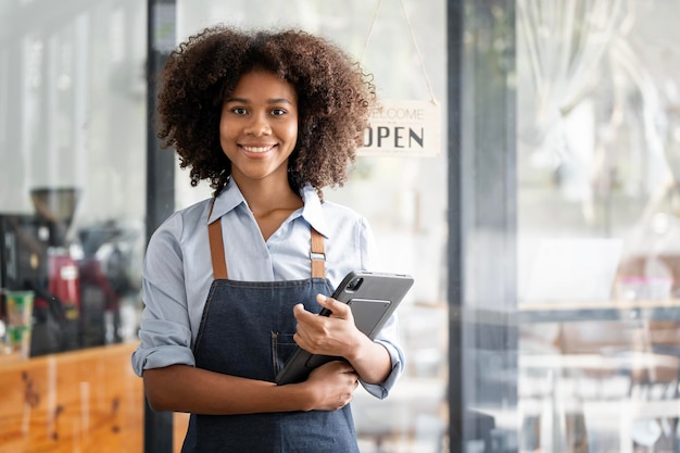 Porträt einer lächelnden jungen afroamerikanischen Barista, die ein Tablet hält, das in ihrem Geschäft steht