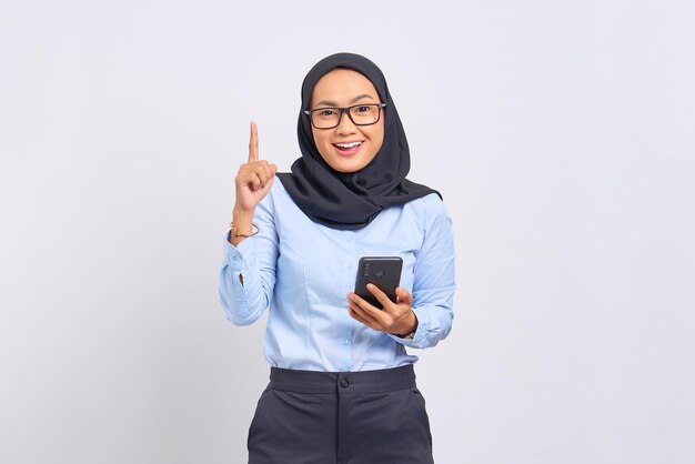 Porträt einer lächelnden jungen Asiatin, die mit dem Finger nach oben zeigt und das Handy isoliert auf weißem Hintergrund hält