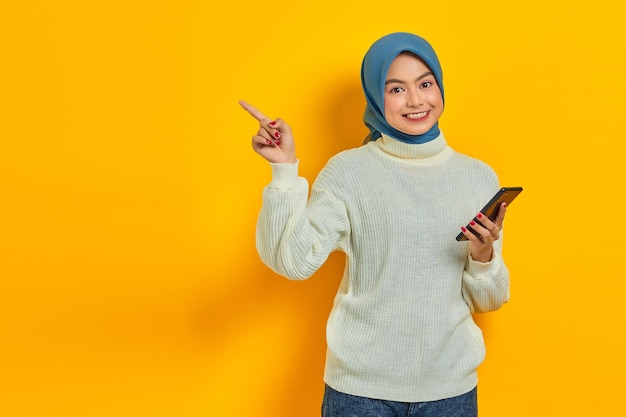 Porträt einer lächelnden jungen asiatischen Frau in weißem Pullover und Hijab, die ein Handy hält, das mit dem Finger zur Seite zeigt, isoliert über gelbem Hintergrund Religiöses Konzept des Menschen islam