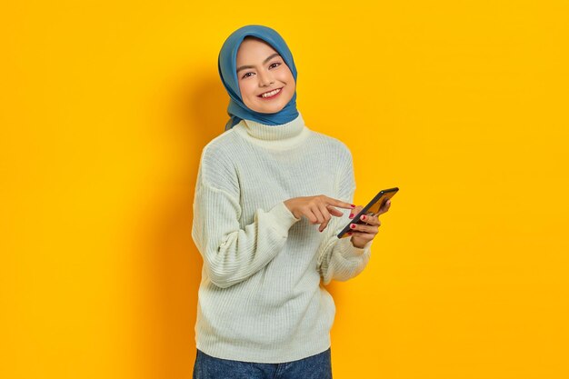 Porträt einer lächelnden jungen asiatischen Frau in weißem Pullover und Hijab, die mit dem Handy mit dem Finger auf das Bildschirmtelefon zeigt, das über gelbem Hintergrund isoliert ist Menschen islam religiöses Konzept