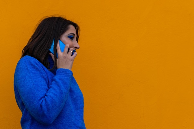 Porträt einer lächelnden jungen brünetten Frau in blauem Kleid und Make-up, die am Telefon auf gelbem Hintergrund spricht