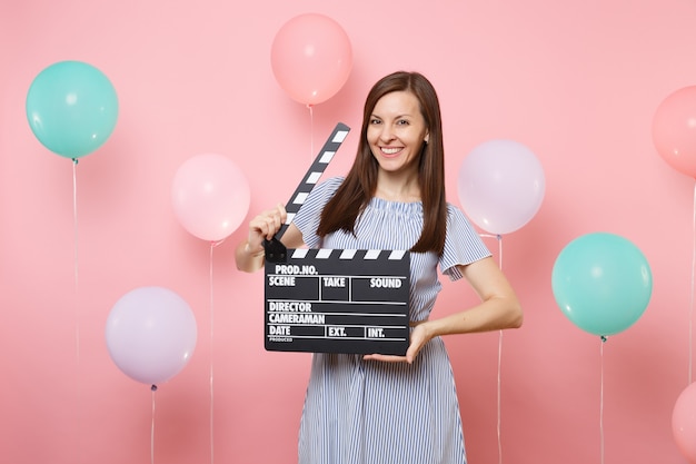 Porträt einer lächelnden jungen Frau, die blaues Kleid trägt und klassische schwarze Filmklappe auf rosafarbenem Hintergrund mit bunten Luftballons hält. Geburtstagsfeier, Konzept der aufrichtigen Emotionen der Menschen.