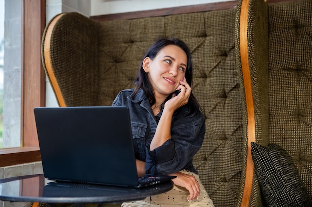 Porträt einer lächelnden jungen Frau, die einen Laptop benutzt und mit dem Mobiltelefon spricht