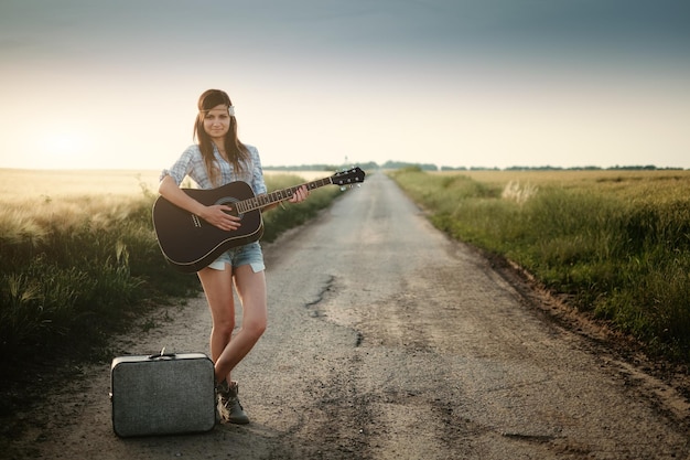 Porträt einer lächelnden jungen Frau, die Gitarre spielt, während sie auf einer Schotterstraße inmitten eines Feldes steht