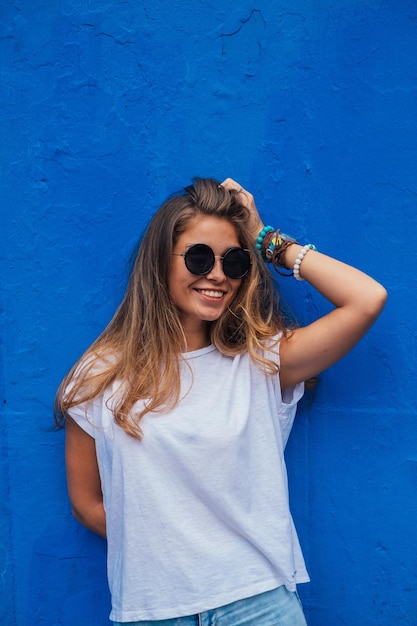 Porträt einer lächelnden jungen Frau mit Sonnenbrille, die vor einer blauen Wand steht