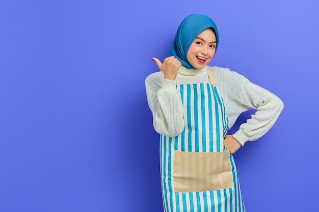 Porträt einer lächelnden jungen Hausfrau in Hijab und Schürze, die auf die Kamera blickt, die mit dem Finger zur Seite zeigt, isoliert auf violettem Hintergrund Menschen Hausfrau muslimischen Lifestyle-Konzept
