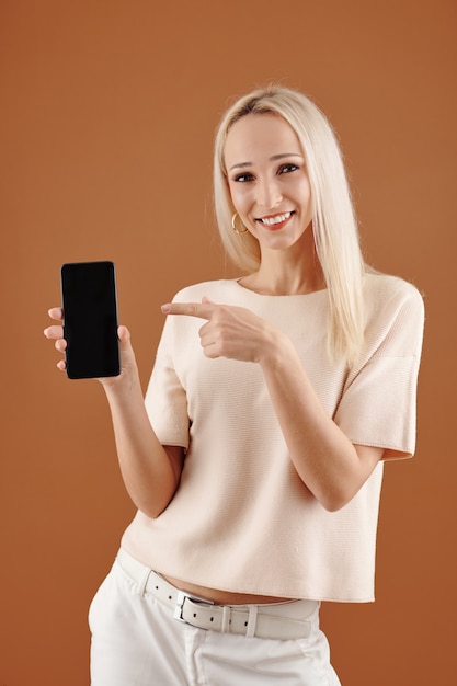 Foto porträt einer lächelnden jungen hübschen frau, die auf den bildschirm des smartphones in der hand zeigt und in die kamera schaut