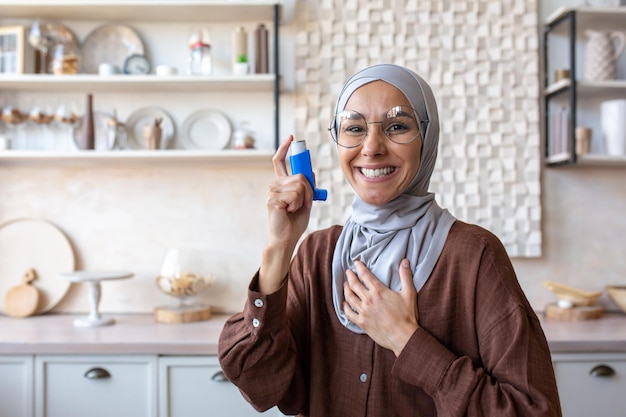 Porträt einer lächelnden jungen muslimischen Frau im Hijab, die zu Hause einen Asthma- und Allergieinhalator in der Hand hält