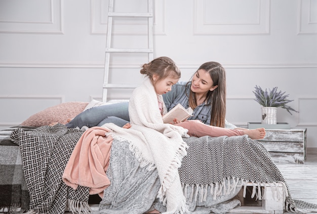 Porträt einer lächelnden jungen niedlichen Mutter und Tochter, die ein liegendes Buch lesen und sich im Bett in einem hellen großen weißen Raum entspannen.