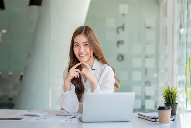 Porträt einer lächelnden jungen schönen asiatischen Geschäftsfrau, die mit einem Laptop sitzt und im Büro in die Kamera blickt