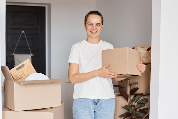 Porträt einer lächelnden kaukasischen Frau mit weißem T-Shirt und Jeans, die mit einer Kartonschachtel steht, die in eine neue Wohnung umzieht und Glück ausdrückt