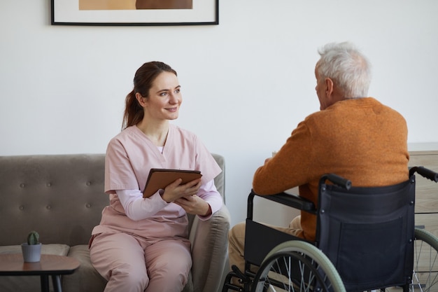 Porträt einer lächelnden Krankenschwester, die mit einem älteren Mann im Rollstuhl spricht und ein digitales Tablet im Altersheim verwendet, Platz kopieren