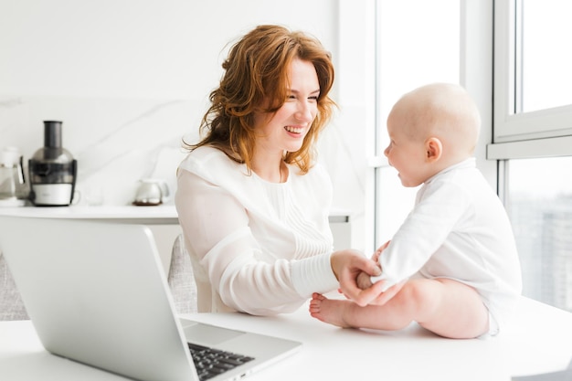 Porträt einer lächelnden Mutter, die ihr fröhliches kleines Baby mit einem Laptop in der Nähe sitzt und glücklich betrachtet