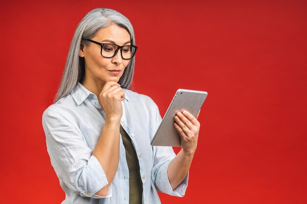 Porträt einer lächelnden, schönen asiatischen Seniorin im Alter von reifer Frau, die Tablet-Computer verwendet, die über rotem Hintergrund isoliert ist Kopieren Sie Platz für Text