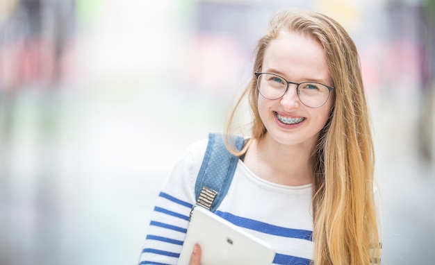 Porträt einer lächelnden schönen Jugendlichen mit Zahnspangen. Junges Schulmädchen mit Schultasche und Tablet-Gerät.