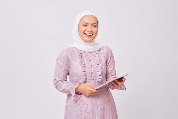 Porträt einer lächelnden schönen jungen asiatischen muslimischen Frau mit Hijab und lila Kleid, die ein digitales Tablet isoliert auf weißem Studiohintergrund hält