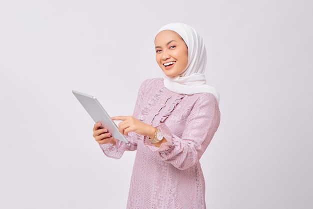 Porträt einer lächelnden schönen jungen asiatischen muslimischen Frau mit Hijab und lila Kleid, die ein digitales Tablet isoliert auf weißem Studiohintergrund hält