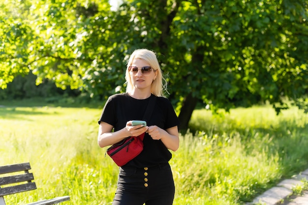 Porträt einer lächelnden, schönen jungen Frau aus nächster Nähe mit einem Mobiltelefon vor dem Hintergrund eines sommergrünen Parks.