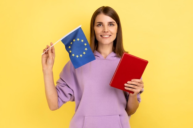 Foto porträt einer lächelnden schönen studentin, die bücher und die flagge der europäischen union hält, bildung im ausland, lila hoodie trägt. studioaufnahme im innenbereich isoliert auf gelbem hintergrund.