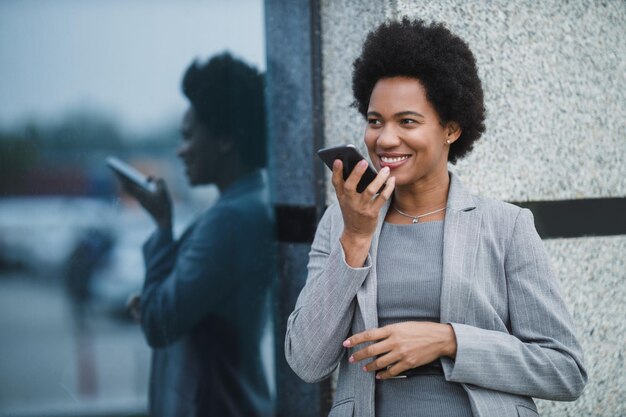 Porträt einer lächelnden schwarzen Geschäftsfrau, die während einer kurzen Pause vor einem Firmengebäude eine Sprachnachricht auf einem Smartphone spricht.