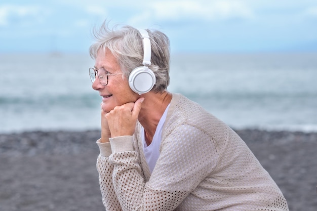 Porträt einer lächelnden Seniorin mit Brille und Kopfhörern, die am Strand Musik hört und Musikfreiheit und Urlaub genießt Horizont über Wasser kopieren Raum