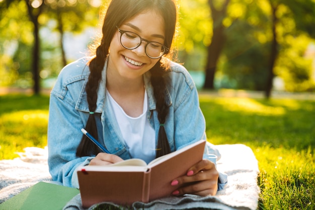 Porträt einer lächelnden tragenden Brille des jungen Studentenmädchens, die draußen im Naturpark sitzt, der Anmerkungen Lesebuch schreibt.