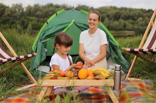 Porträt einer lächelnden zufriedenen frau und eines kleinen mädchens, die auf einer decke in der nähe des zeltes sitzen und eine picknick-tochter mit pfirsich haben, fühlt sich hungrig, dass die familie über nacht in der nähe des flusses bleibt