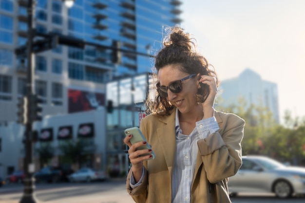 Porträt einer Latina-Frau mit Sonnenbrille, die beim Betrachten des Handys, der Stadt im Hintergrund, des selektiven Fokus und des Kopierraums lächelt