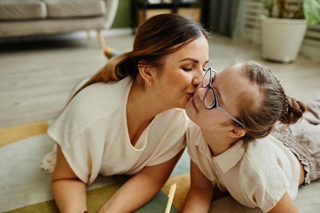 Porträt einer liebevollen Mutter, die Tochter mit Down-Syndrom küsst, während sie zusammen auf dem Boden liegt, nicht traditionelle Familie