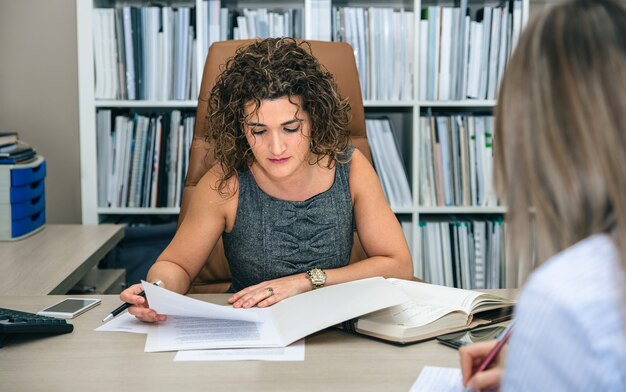 Porträt einer lockigen Geschäftsfrau, die im Büro mit Dokumenten arbeitet, während die blonde Sekretärin Notizen macht