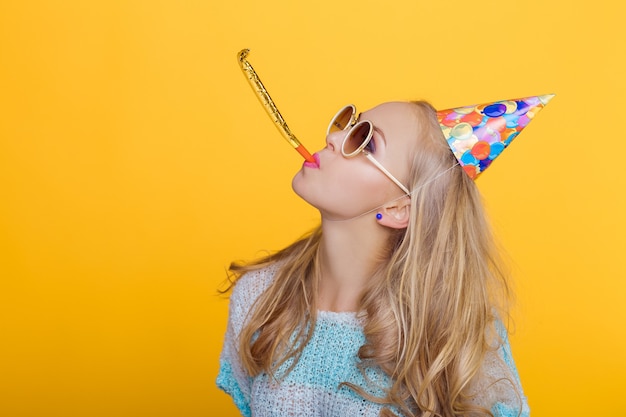 Porträt einer lustigen blonden frau in geburtstagshut und blauem hemd auf gelbem hintergrund, feier und party