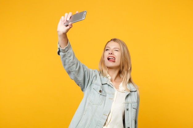 Porträt einer lustigen jungen Frau in Denim-Freizeitkleidung, die Zunge zeigt und Selfie-Aufnahmen auf dem Handy macht, isoliert auf gelb-orangefarbenem Hintergrund im Studio. People-Lifestyle-Konzept. Mock-up-Kopienbereich.