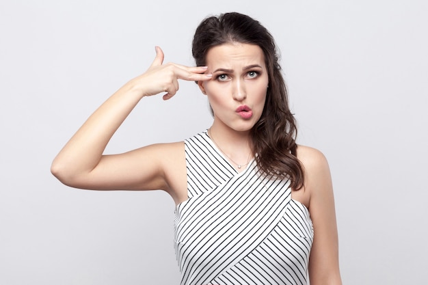 Porträt einer lustigen, verrückten, schönen jungen Brunettefrau mit Make-up und gestreiftem Kleid, die mit Pistolengeste steht und wegschaut. Indoor-Studioaufnahme, auf grauem Hintergrund isoliert.