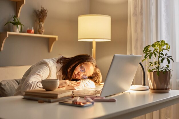 Porträt einer müden, schläfrigen kaukasischen Frau mit braunen Haaren, die am Tisch sitzt und abends zu Hause am Laptop arbeitet und überarbeitet schläft und sich an ihren Schreibtisch lehnt