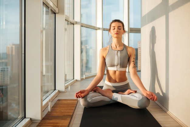 Porträt einer nachdenklichen Dame in sportlichem Oberteil und Leggings, die in Lotus-Pose sitzt und zu Hause auf einer Yogamatte meditiert, mit großen, schönen Fenstern im Hintergrund