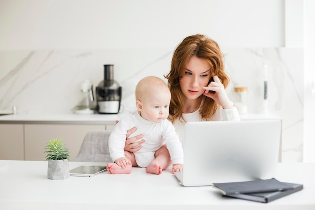 Porträt einer nachdenklichen Geschäftsfrau, die am Tisch sitzt und am Laptop arbeitet, während sie ihr süßes kleines Baby hält