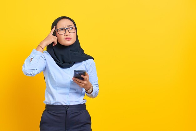 Porträt einer nachdenklichen jungen Asiatin, die ein Mobiltelefon benutzt und auf gelbem Hintergrund nach oben schaut