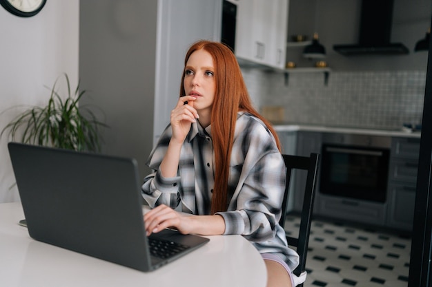 Porträt einer nachdenklichen jungen Frau, die mit einem Laptop am Tisch sitzt und nachdenklich im Kücheninterieur wegschaut. Nachdenkliche rothaarige Studentin, die vom Heimbüro aus auf dem Laptop lernt