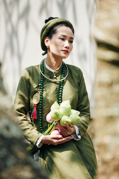 Porträt einer nachdenklichen jungen Frau mit Lotusblumen, die im traditionellen Ao Dai-Kleid im Freien sitzt