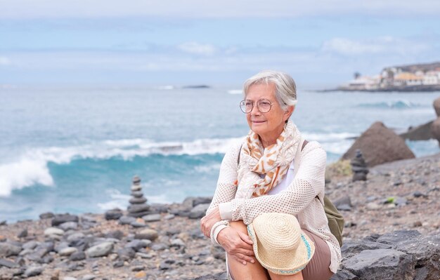 Porträt einer nachdenklichen kaukasischen Frau, die am Strand sitzt und einen Rucksack hält Lächelnde ältere Dame, die Urlaub oder Ruhestand genießt Horizont über dem Wasserkopierraum