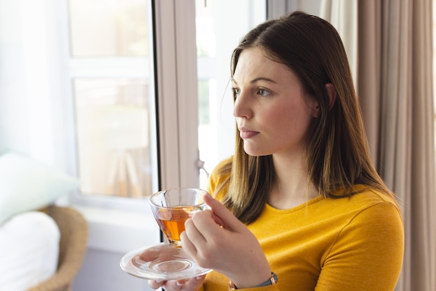 Porträt einer nachdenklichen kaukasischen Frau, die eine Teetasse am Fenster im Wohnzimmer zu Hause hält