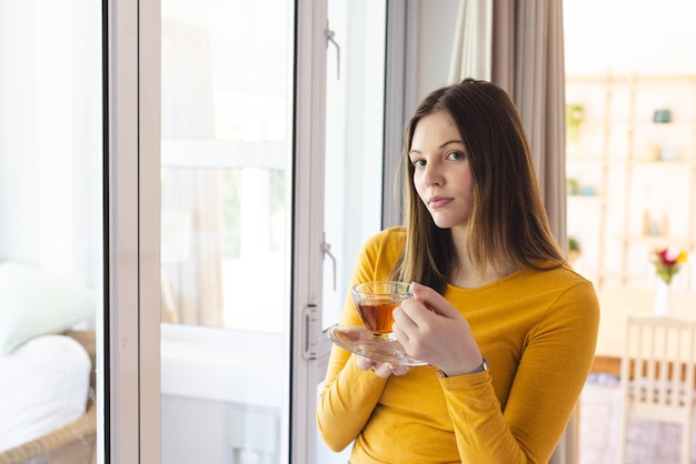 Porträt einer nachdenklichen kaukasischen Frau, die eine Teetasse am Fenster im Wohnzimmer zu Hause hält