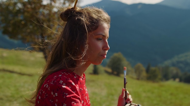 Porträt einer nachdenklichen Künstlerin, die mit Pinsel auf die Natur malt Nahaufnahme eines kreativen Mädchens, das an Kunstwerken arbeitet und im Sommer in den Bergen steht, sonniger Tag. Inspirierter Maler, der ein Bild auf einer grünen Wiese schafft